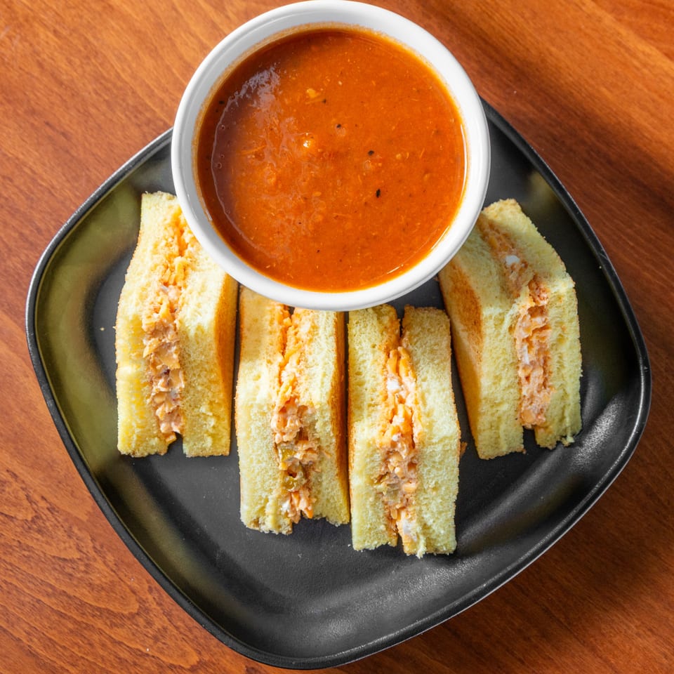 Four deli sandwiches with creamy filling on a black plate, served with a bowl of tomato soup.