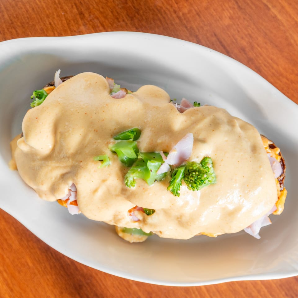 A deli sandwich covered in a creamy sauce and topped with broccoli florets in a white dish.