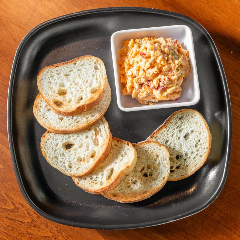 Pimento cheese dip in a white bowl, surrounded by toasted bread slices on a black plate.