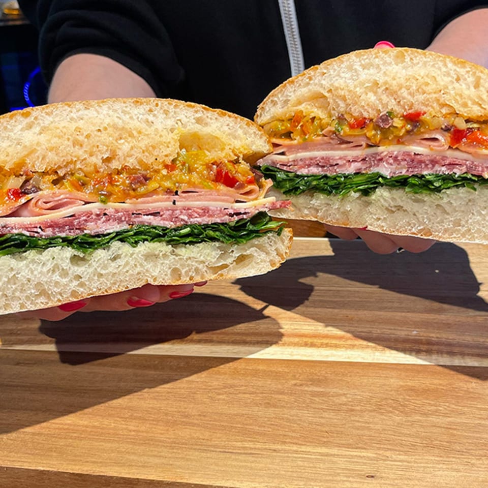 Hands holding a large deli sandwich, cut in half, revealing layers of meat, cheese, and greens.