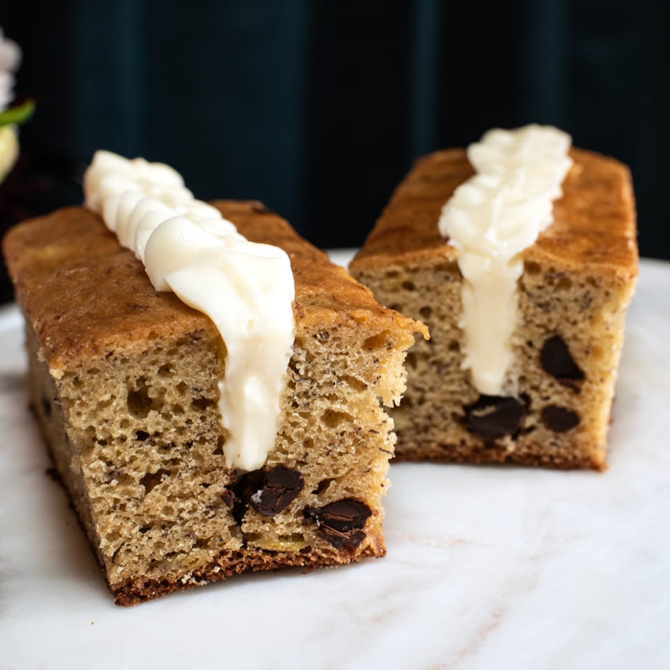 Two slices of chocolate chip banana bread with cream cheese frosting on a marble surface.